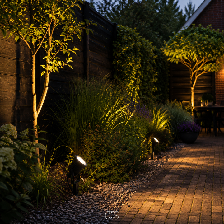 Meerdere tuinspots verlichten in de avond planten, bomen en grind langs een klinkerpad, met een terras op de achtergrond. 4