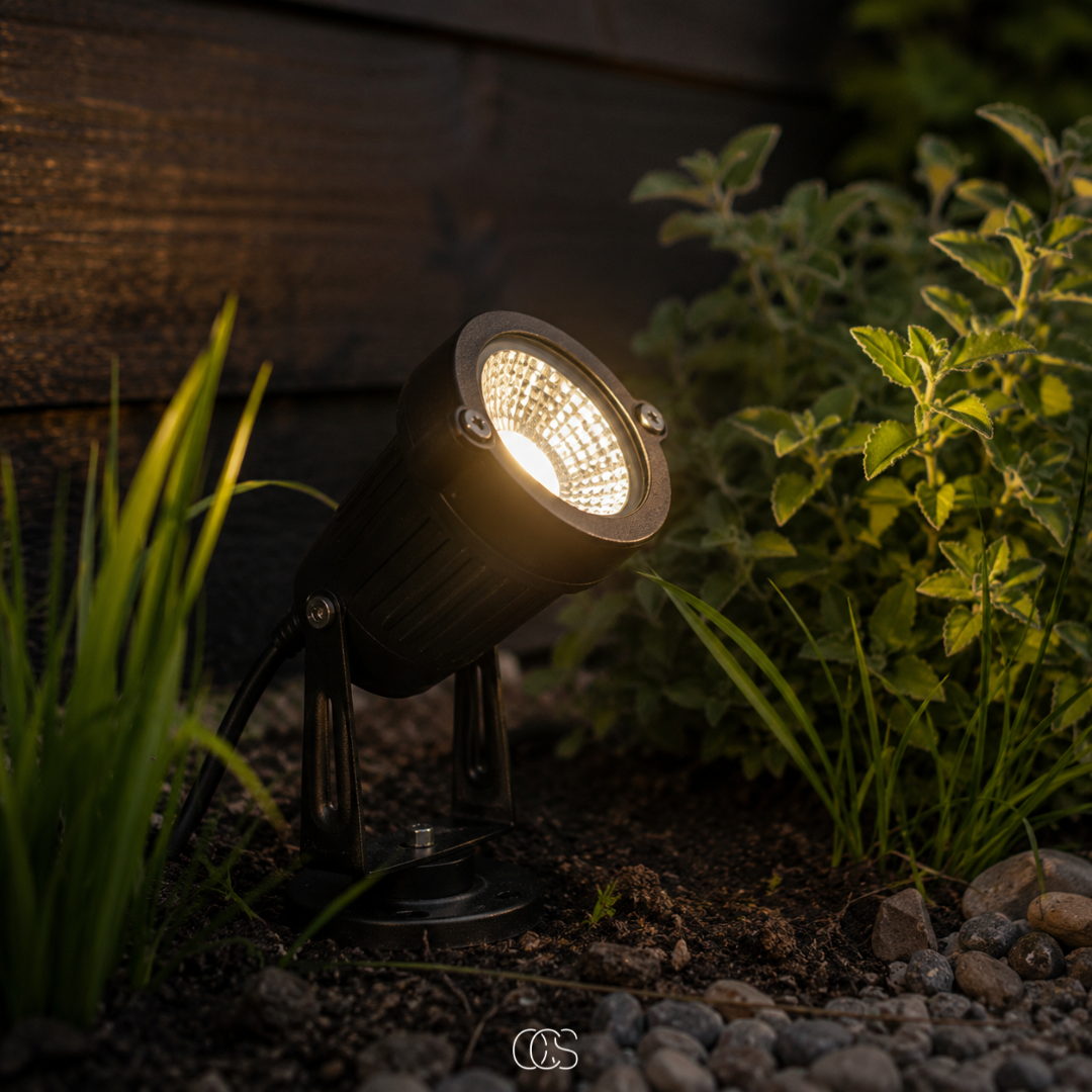 Zwarte tuinspot brandt met warm licht in de grond tussen planten en keitjes, voor een donkere houten schutting in de schemering. 1