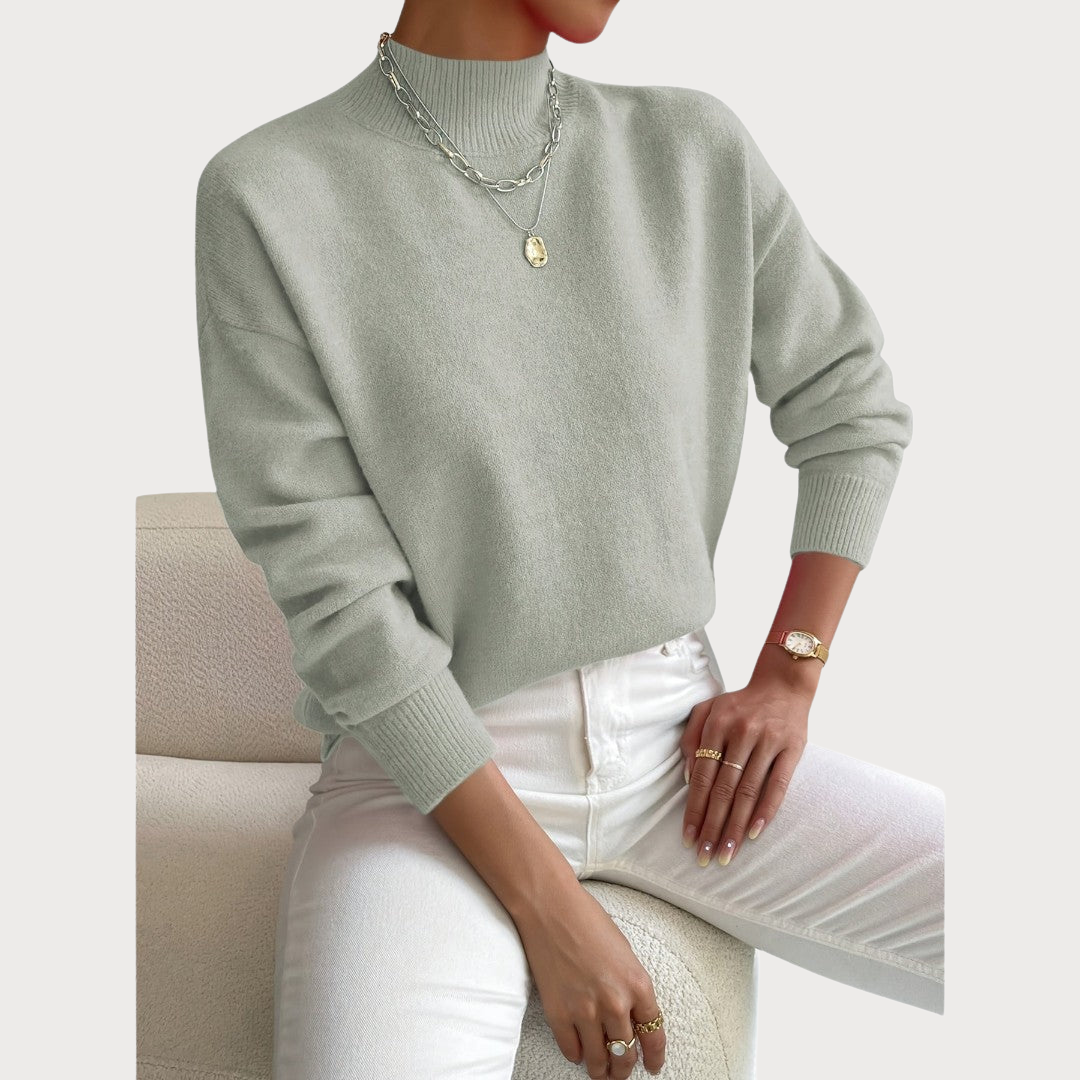Model draagt de trui in saliegroen met ribkraag en manchetten, gecombineerd met witte broek en gouden sieraden. 2