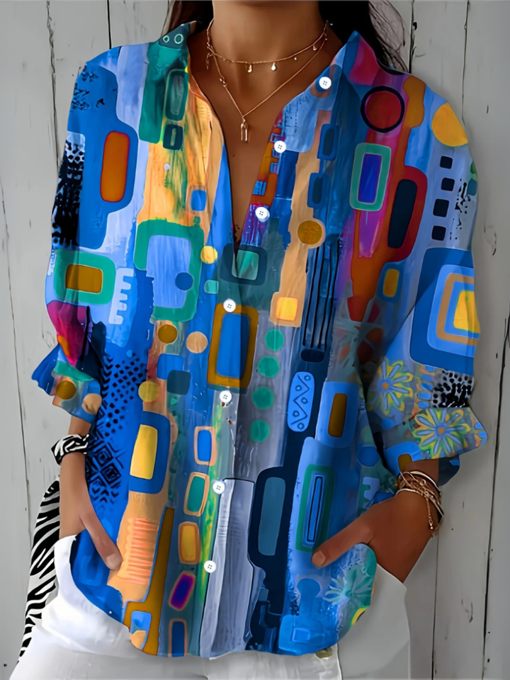 Close-up van de blauwe blouse met geometrische vormen en bloemmotieven op de mouwen, V-hals en knoopsluiting goed zichtbaar. 6