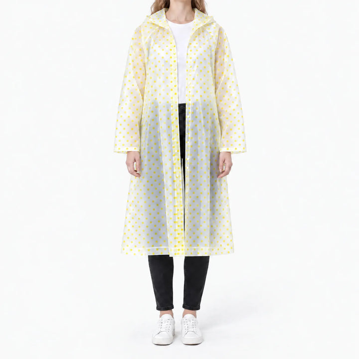 Vrouw in een doorzichtige regenjas met gele stippen en capuchon, open gedragen over wit shirt, zwarte broek en witte sneakers. 11