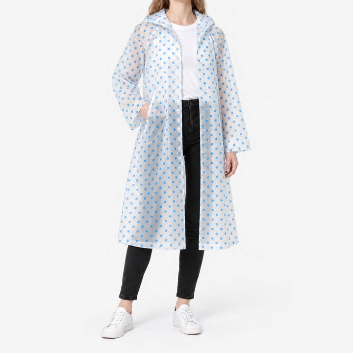 Vrouw draagt een doorzichtige, openstaande regenjas met blauwe stippen over wit shirt en zwarte broek, met een hand in de zak. 2