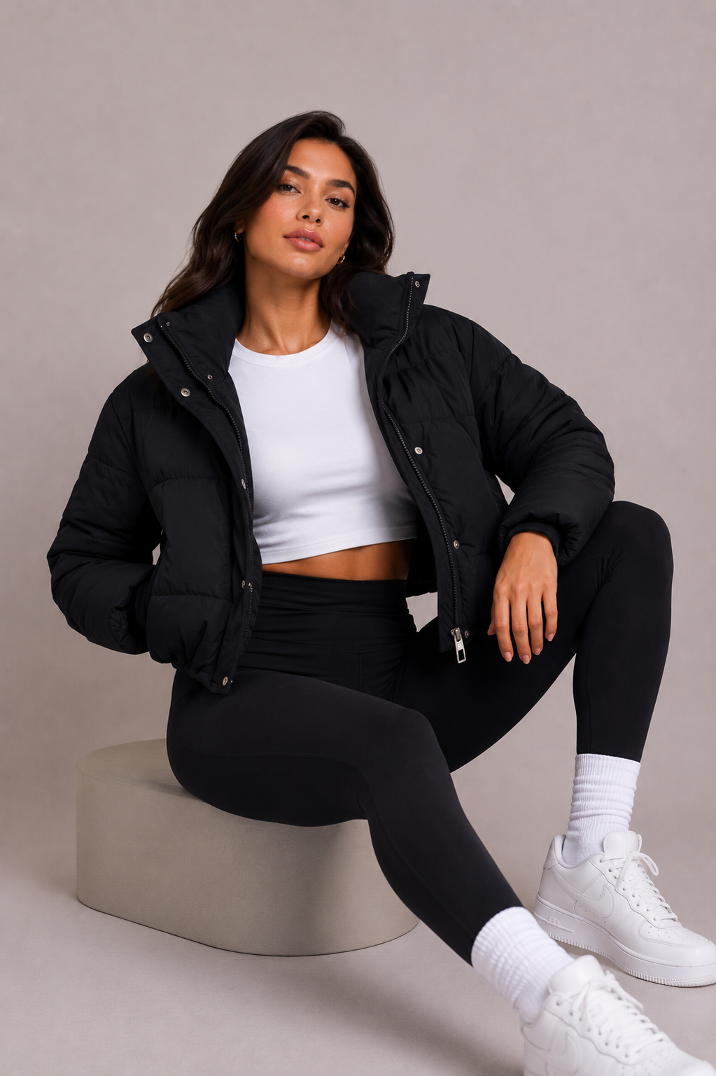Zittend model draagt de korte zwarte pufferjack open over een wit topje, met legging en witte sneakers, in een studiosetting. 6