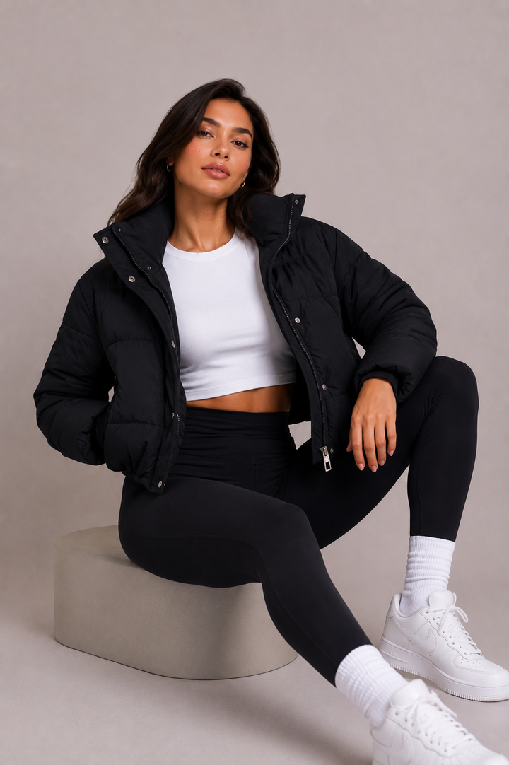 Zittend model draagt de korte zwarte pufferjack open over een wit topje, met legging en witte sneakers, in een studiosetting. 6