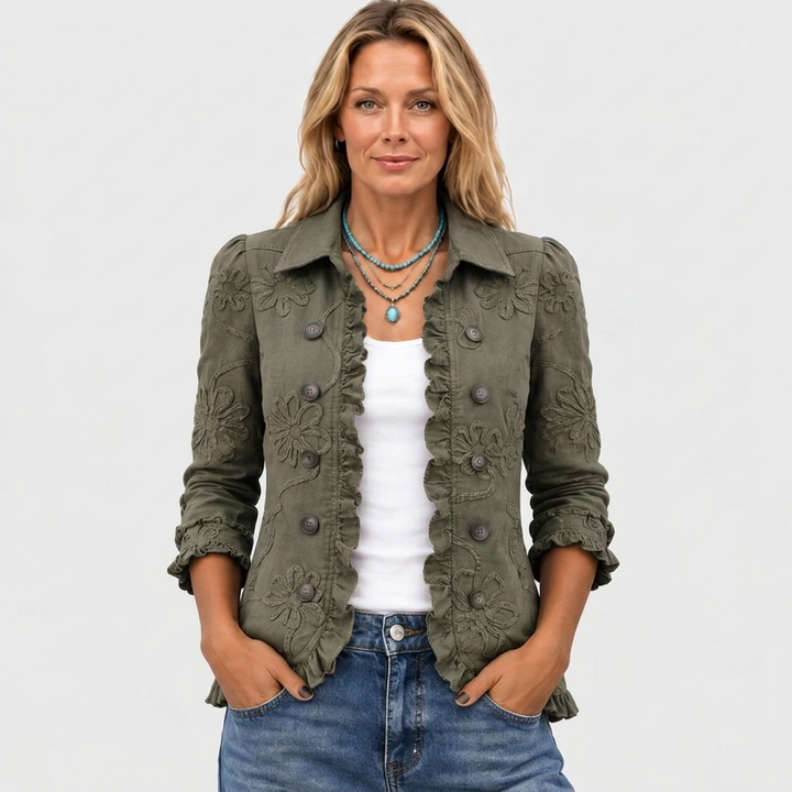 Model draagt het olijfgroene jasje open van voren met witte top, spijkerbroek en turquoise kettingen, handen in de zakken. 2