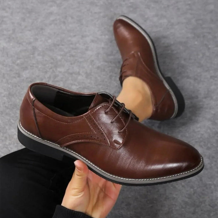 Donkerbruine nette schoen in de hand gehouden boven een grijs tapijt, met de tweede schoen op de achtergrond. 2