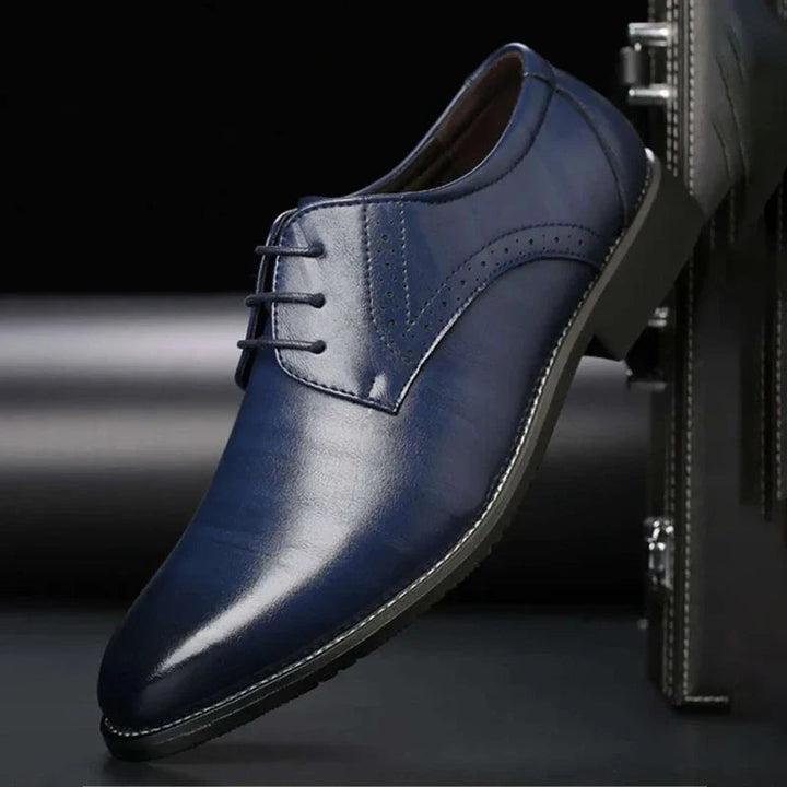 Glanzende donkerblauwe nette schoen met gesloten vetering en brogue-perforatie, schuin geplaatst tegen een donkere koffer. 5