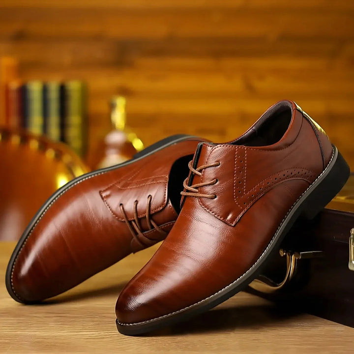 Twee glanzende cognacbruine nette veterschoenen schuin tegen elkaar op een houten tafel, met brogue-perforatie en witte stikselrand. 1