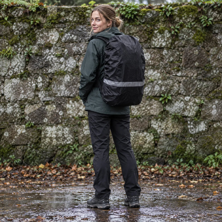 Vrouw in regenjas draagt een zwarte rugzak met regenhoes met reflecterende streep, staand op een natte weg bij een mossige stenen muur. 1