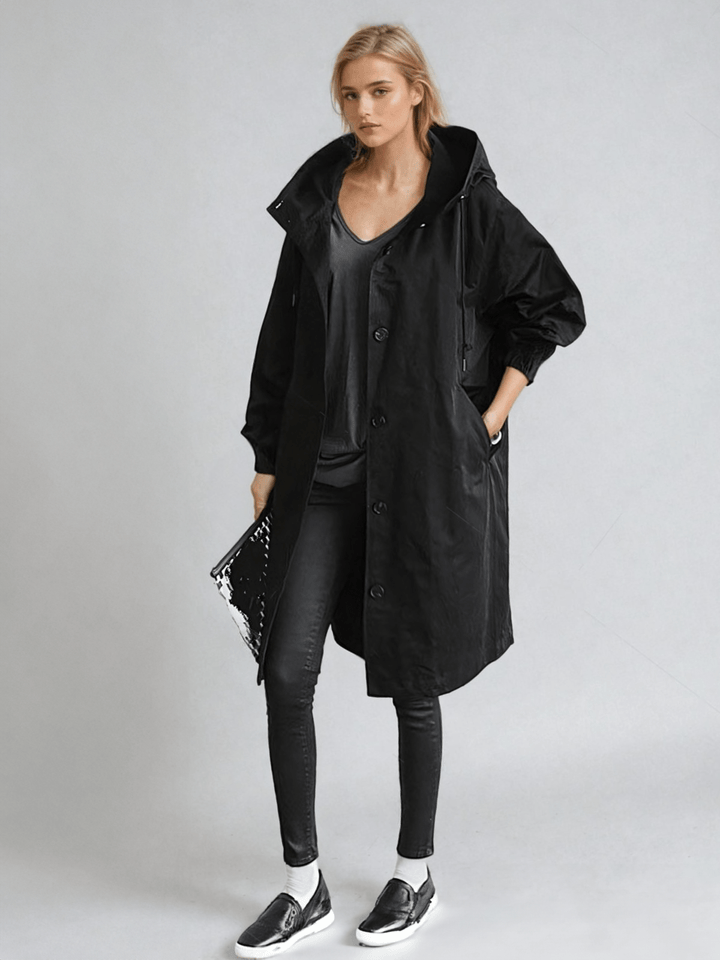 Model in de zwarte trenchcoat met capuchon, gecombineerd met zwarte skinny broek, sneakers en een glanzende clutch onder de arm. 2