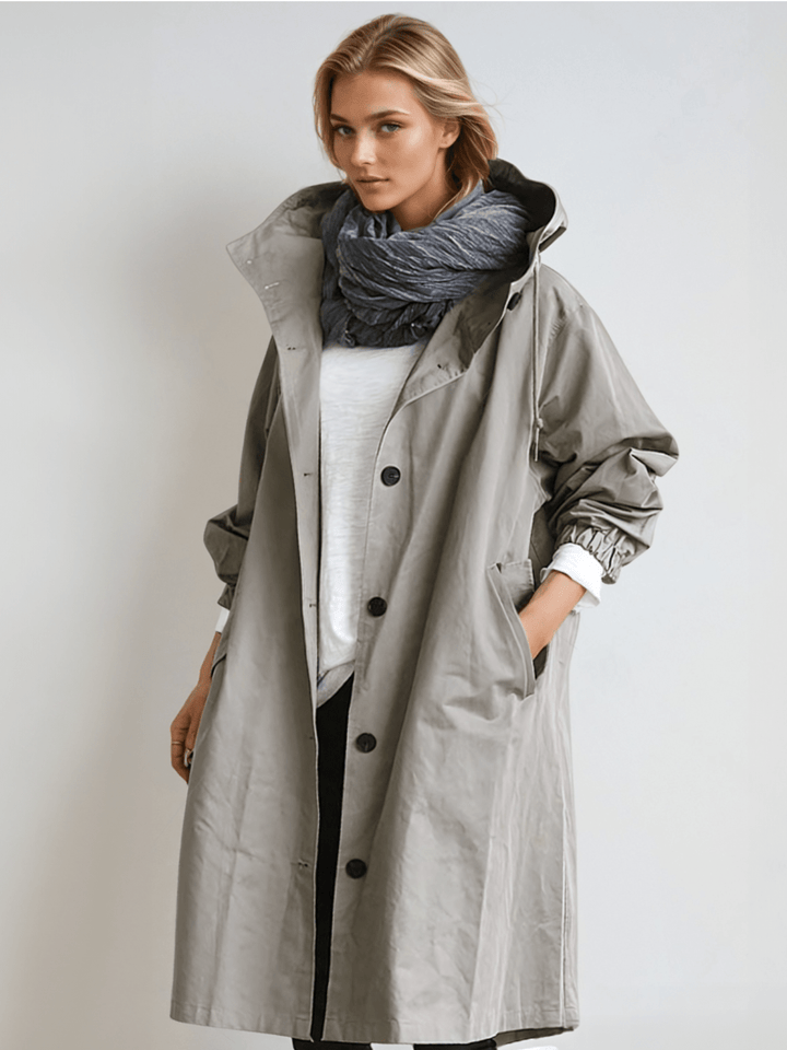 Model draagt de grijze trenchcoat met capuchon open over een wit shirt en donkere sjaal, met een hand in de zak. 1