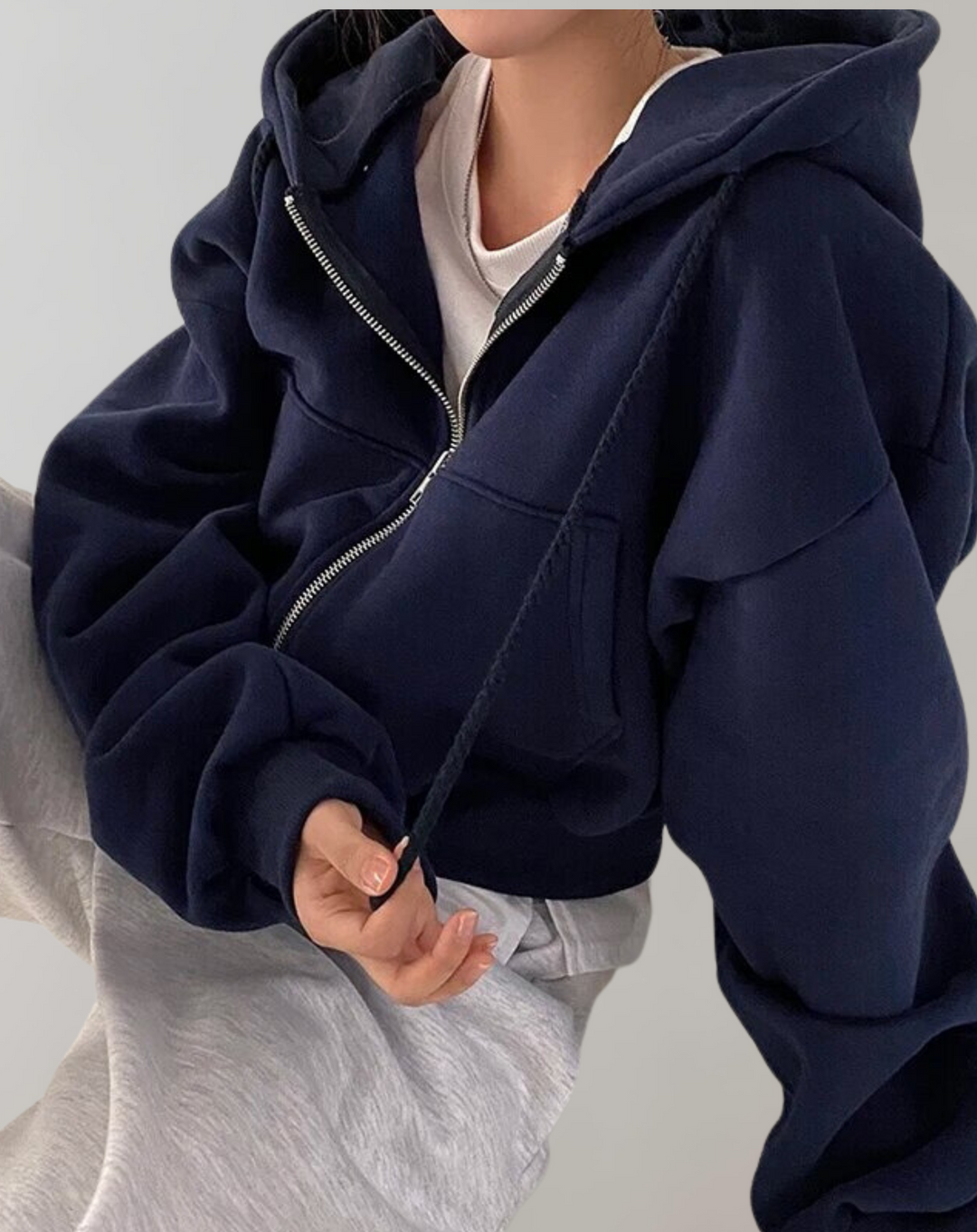 Model draagt de donkerblauwe hoodie met open rits over een wit shirt en lichtgrijze broek, een hand houdt het trekkoord vast. 3