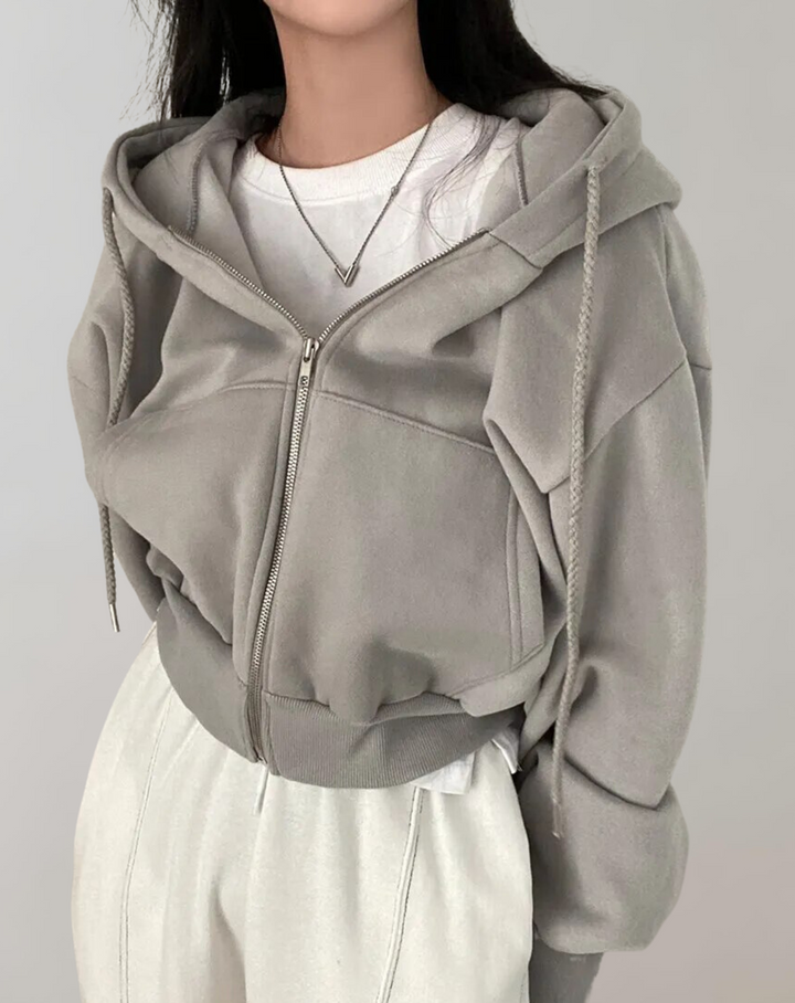 Frontaal beeld van de grijze hoodie met dichte rits en geribde onderrand, gedragen met een wit shirt, ketting en witte broek. 4