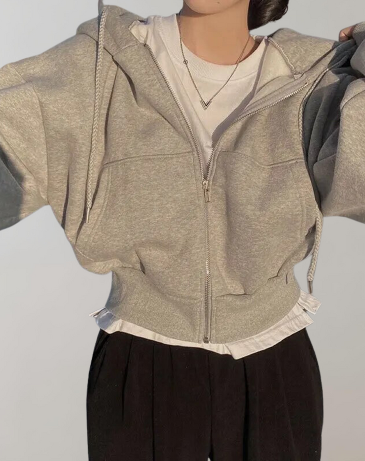 Model draagt de lichtgrijze hoodie met rits over een wit shirt en zwarte broek, met geribde zoom en vlechtkoorden. 5