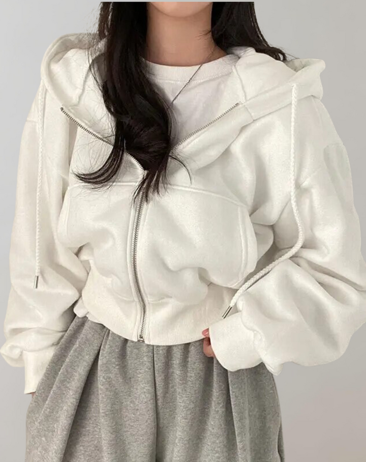 Model draagt de witte hoodie met rits half open over een wit shirt en grijze joggingbroek, met trekkoorden langs de capuchon. 1