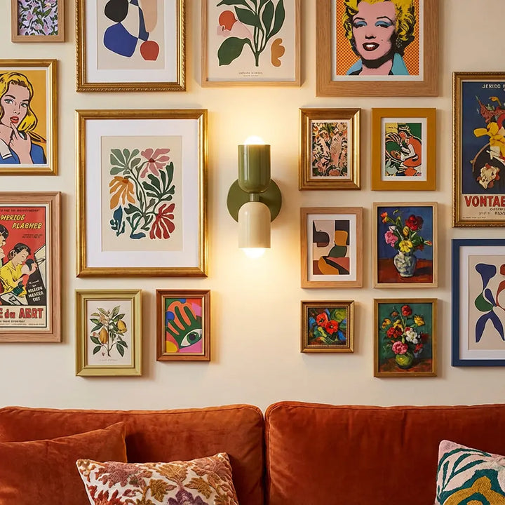Brandende wandlamp in olijfgroen en crème midden in een galerijwand met kleurrijke prints, boven een roestkleurige bank. 6