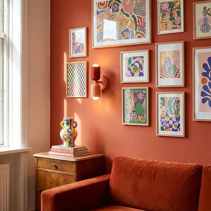 Wandlamp in rood en perzik tussen kleurrijke ingelijste prints op een terracotta muur, boven een bank en een beschilderde vaas. 5