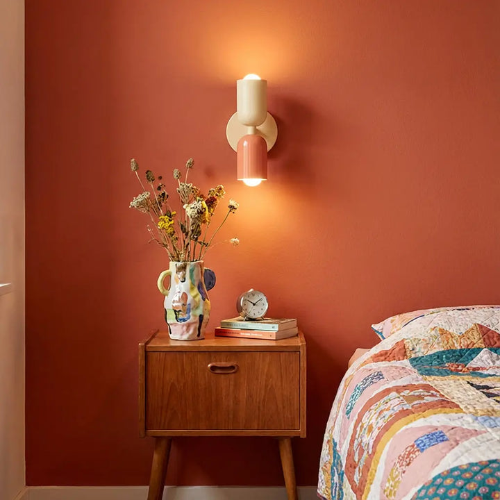 Brandende wandlamp in crème en zalmroze boven een houten nachtkastje, tegen een terracotta muur naast een quilt-dekbed. 1