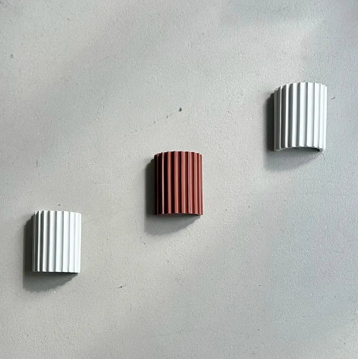 Drie geribbelde wandlampen in wit, terracotta en lichtgrijs, diagonaal verspreid op een grijze muur 13
