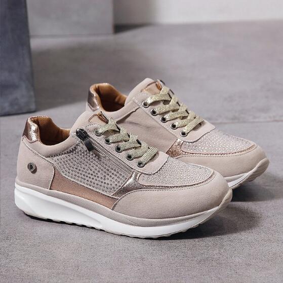Twee beige sneakers op een grijze vloer, met gouden veters, een rits en fijne strasssteentjes op het bovenwerk. 2