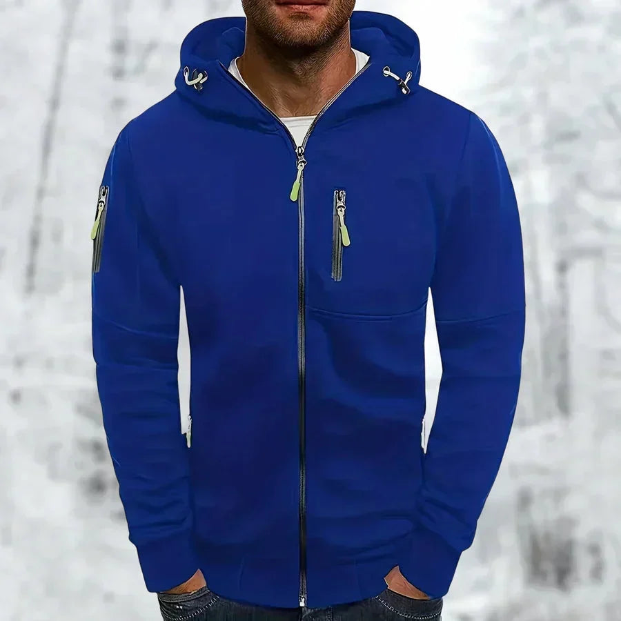 Model in kobaltblauwe hoodie met rits, neongroene ritstrekkers en borstzak, voorkant, tegen een besneeuwde bosachtergrond. 1