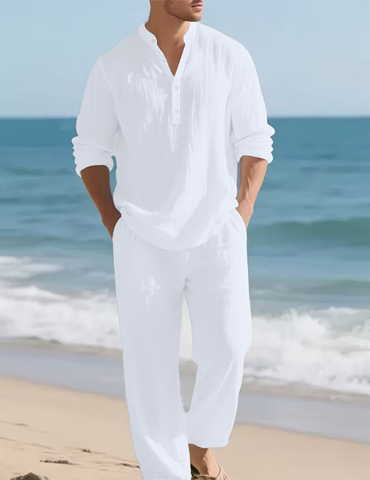 Model in de witte set met opgerolde mouwen en handen in de zakken, wandelend langs de zee op het strand. 2