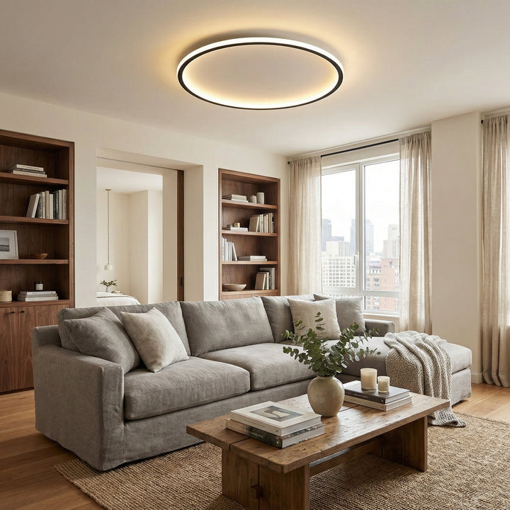 Ringvormige plafondlamp met warm licht boven een grijze hoekbank, houten salontafel en boekenkasten in een woonkamer. 4