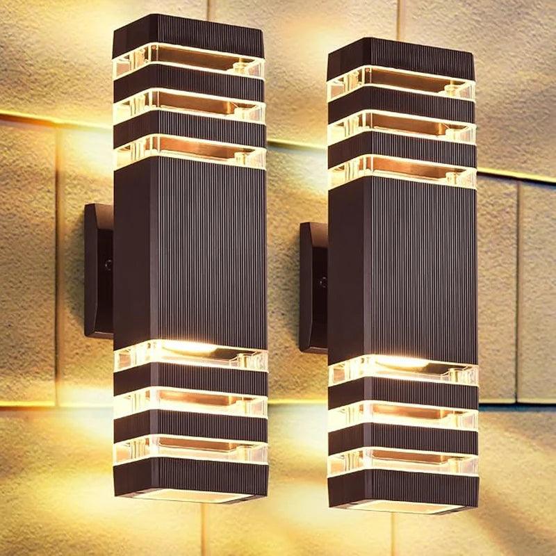 Twee donkerbruine, rechthoekige wandlampen met ribbelstructuur en heldere strepen, die warm licht omhoog en omlaag werpen op een stenen gevel. 1