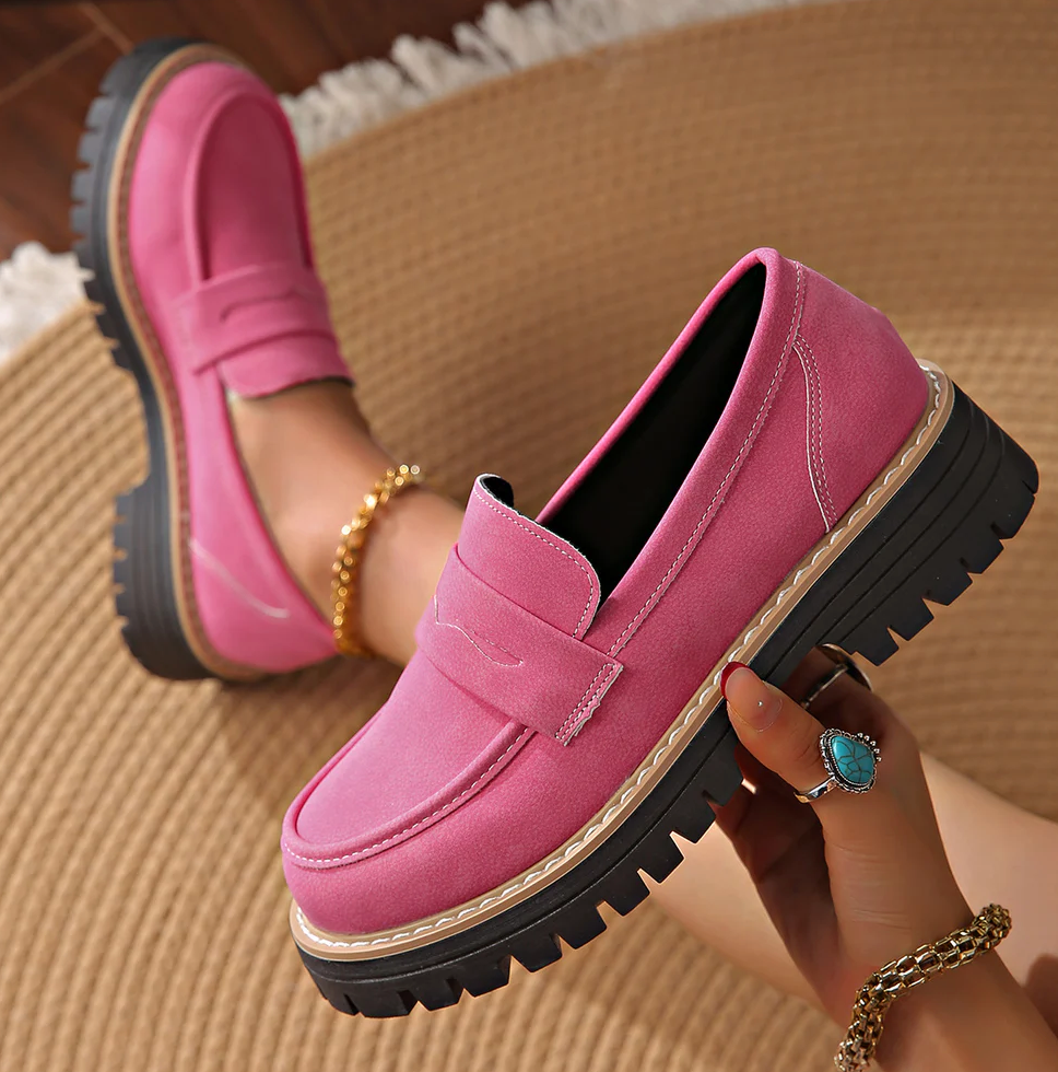 Roze loafers met witte stiksels en dikke zwarte zool, door een model schuin vastgehouden op een rieten ondergrond. 18