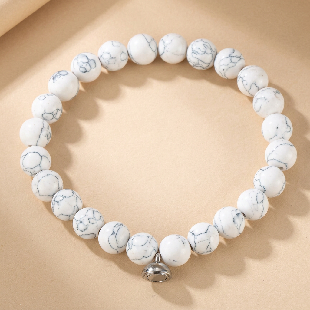Armband van witte kralen met grijze adering en zilverkleurige magneetsluiting, liggend op een beige ondergrond 3