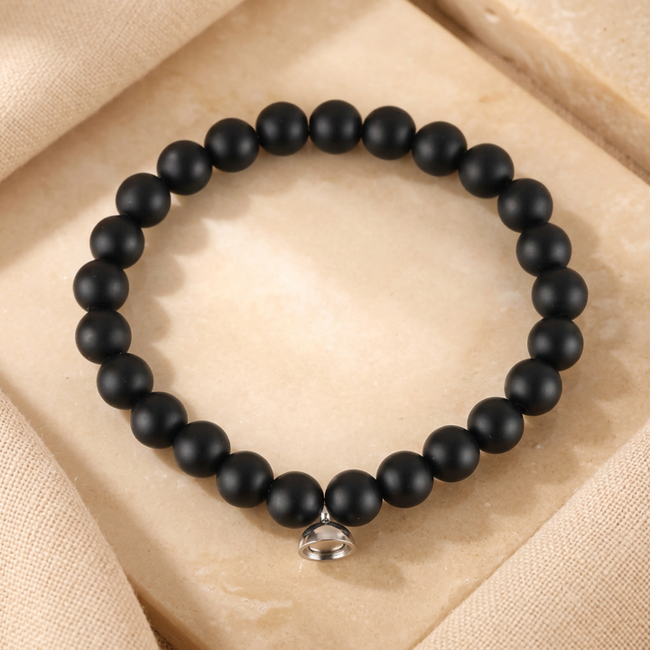 Armband van matzwarte kralen met klein zilverkleurig magneetsluitingsbedeltje, rustend op een beige steen 2