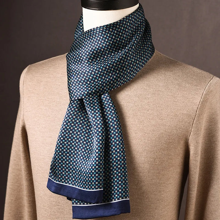 Blauwe zijden sjaal met klein blokjesmotief in turquoise en wit en smalle blauwe rand, om een paspop met beige trui gedrapeerd. 24