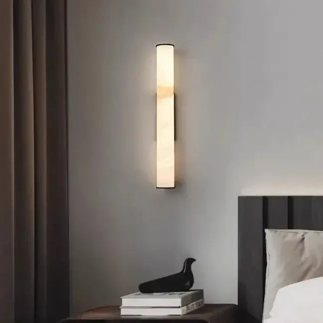 Slanke verlichte wandlamp boven een nachtkastje met boeken en een zwart vogelbeeldje, naast een donker hoofdeinde en gordijn. 14