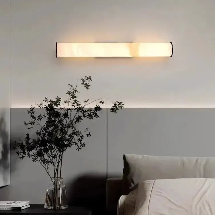 Horizontaal gemonteerde marmeren wandlamp boven een bed, met een glazen vaas met donkere takken ervoor. 16