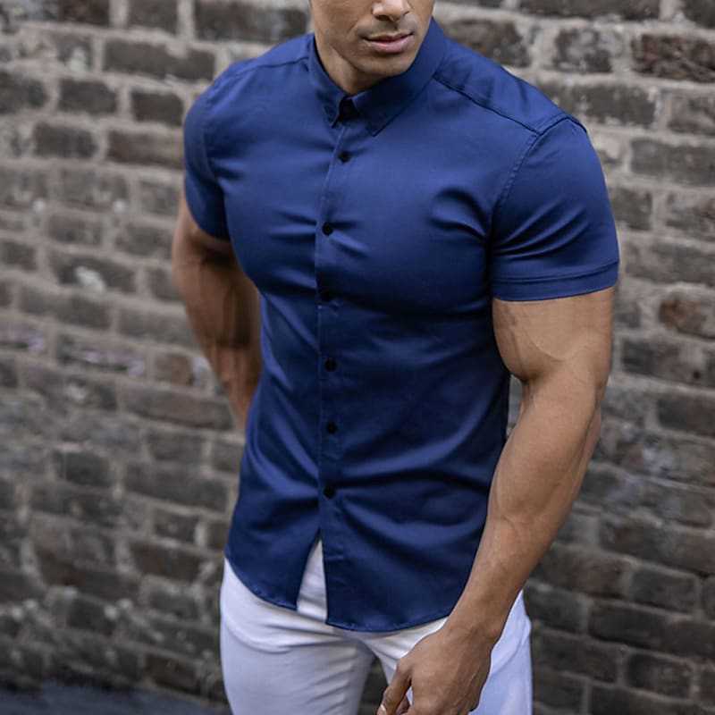 Donkerblauw overhemd met korte mouwen gedragen door een man in een witte broek, voor een bakstenen muur. 3