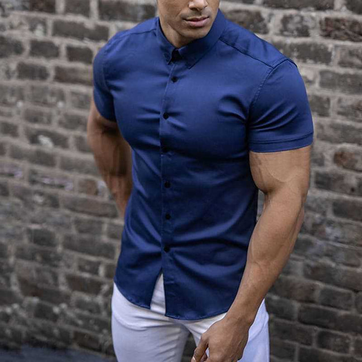 Donkerblauw overhemd met korte mouwen gedragen door een man in een witte broek, voor een bakstenen muur. 3