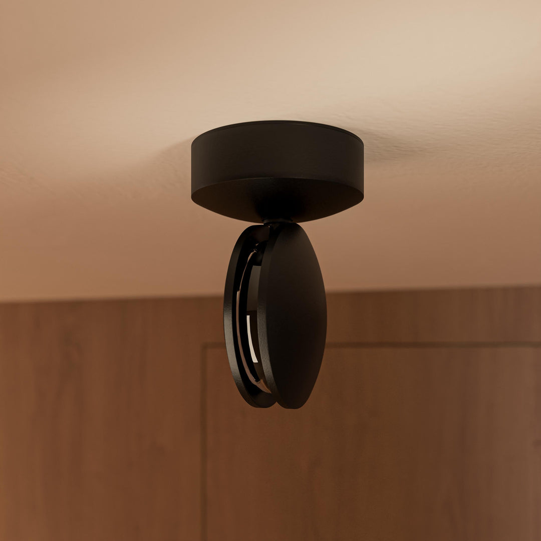 Zwarte plafondlamp van onderen gezien, met de ronde kop gekanteld en de lichtopening zichtbaar voor een houten wand. 13