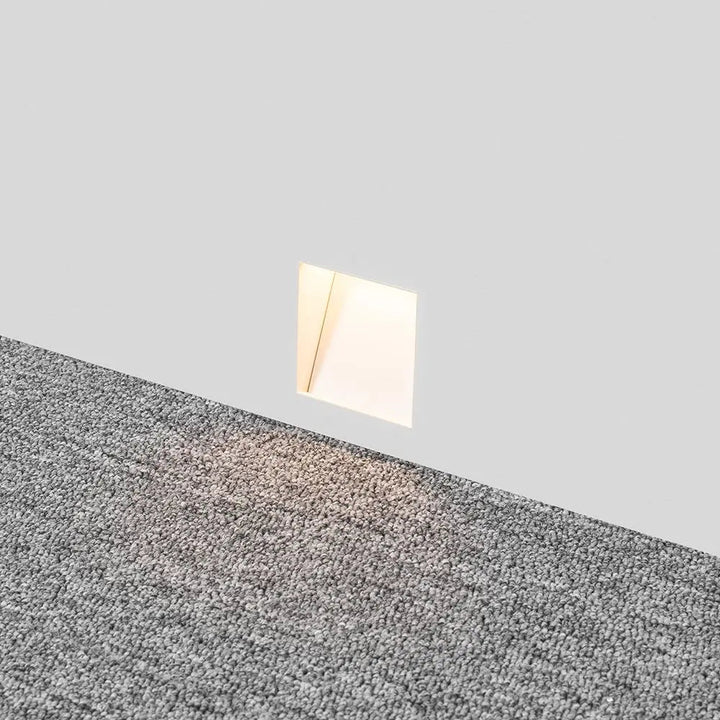 Ingebouwde lamp in een witte wand boven grijs tapijt, met warm lichtschijnsel op de vloer 3