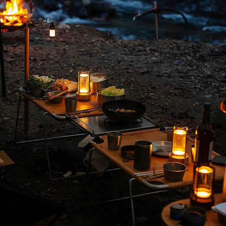 Brandende lampjes op een campingtafel met kookspullen en eten, buiten in het donker bij een kampvuur. 4