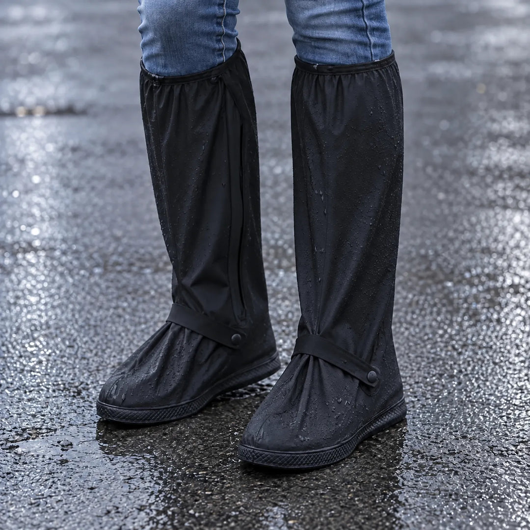 Zwarte regenoverschoenen met waterdruppels, gedragen onder een spijkerbroek op nat asfalt, met bandje en drukknoop bij de voet. 1