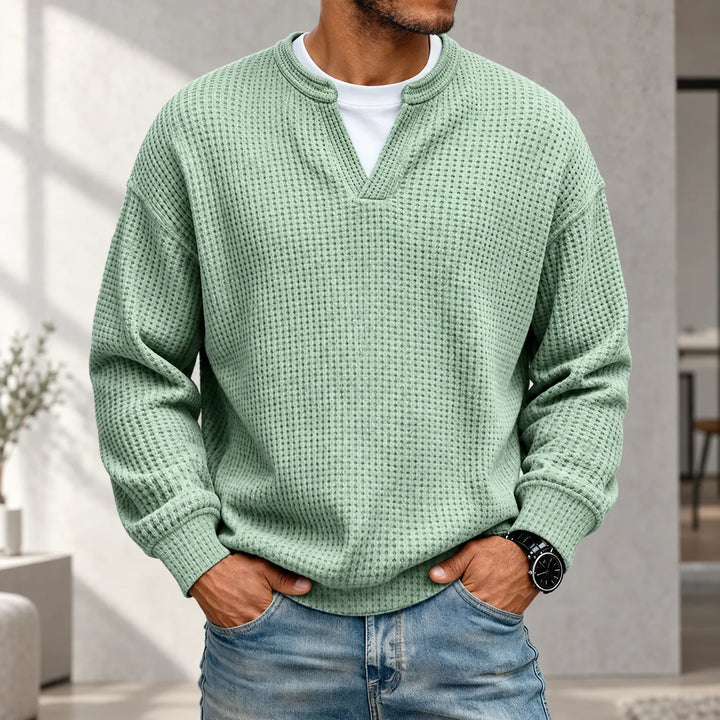 Man draagt de lichtgroene wafeltrui met V-inkeping over een wit shirt, met een spijkerbroek, in een lichte woonkamer. 1