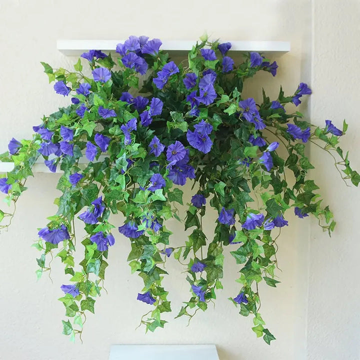 Paarsblauwe kunstbloemen met lange groene hangranken boven een witte wandplank tegen een crèmekleurige muur. 4