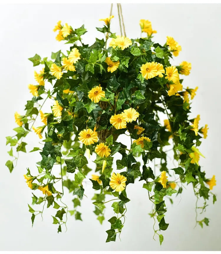 Gele kunstbloemen met groen blad in een hangende mand, opgehangen aan touw tegen een lichte achtergrond. 2