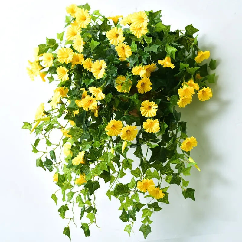 Gele kunstbloemen met groene bladeren en hangende ranken, tegen een witte muur gehangen als wanddecoratie. 5