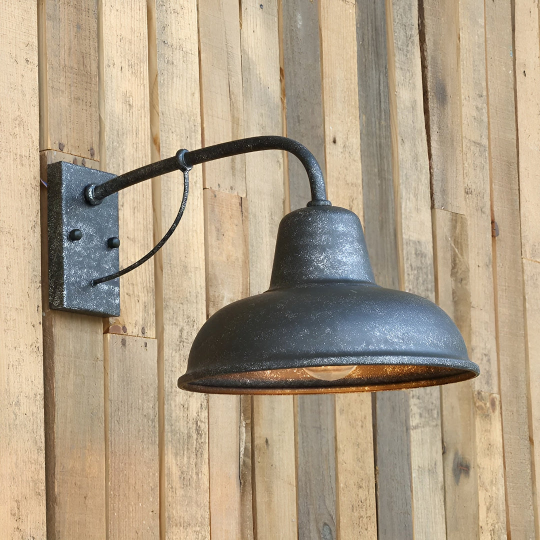 Wandlamp met verweerde, gespikkelde zwartgrijze afwerking bevestigd tegen een wand van houten planken, met brandende lamp onder de kap. 2