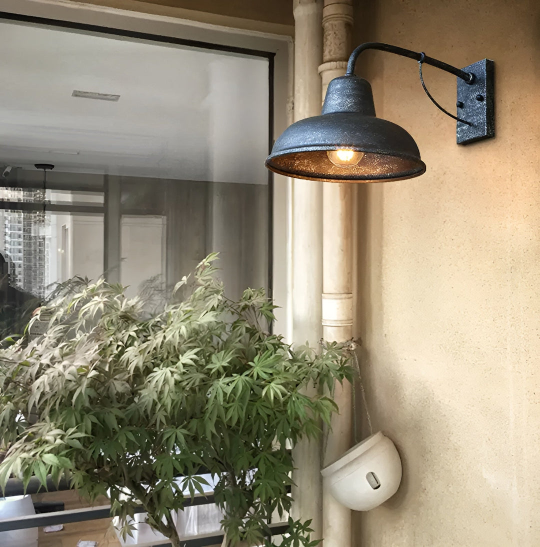 Wandlamp met verweerde afwerking aan een beige buitenmuur naast een raam en een groene plant, met warm brandende lamp. 3