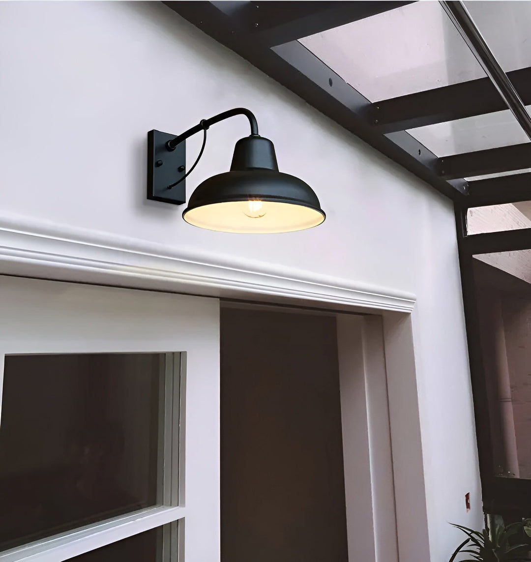 Zwarte wandlamp met witte binnenzijde van de kap, aan een witte gevel boven een deuropening, met brandende gloeilamp. 4