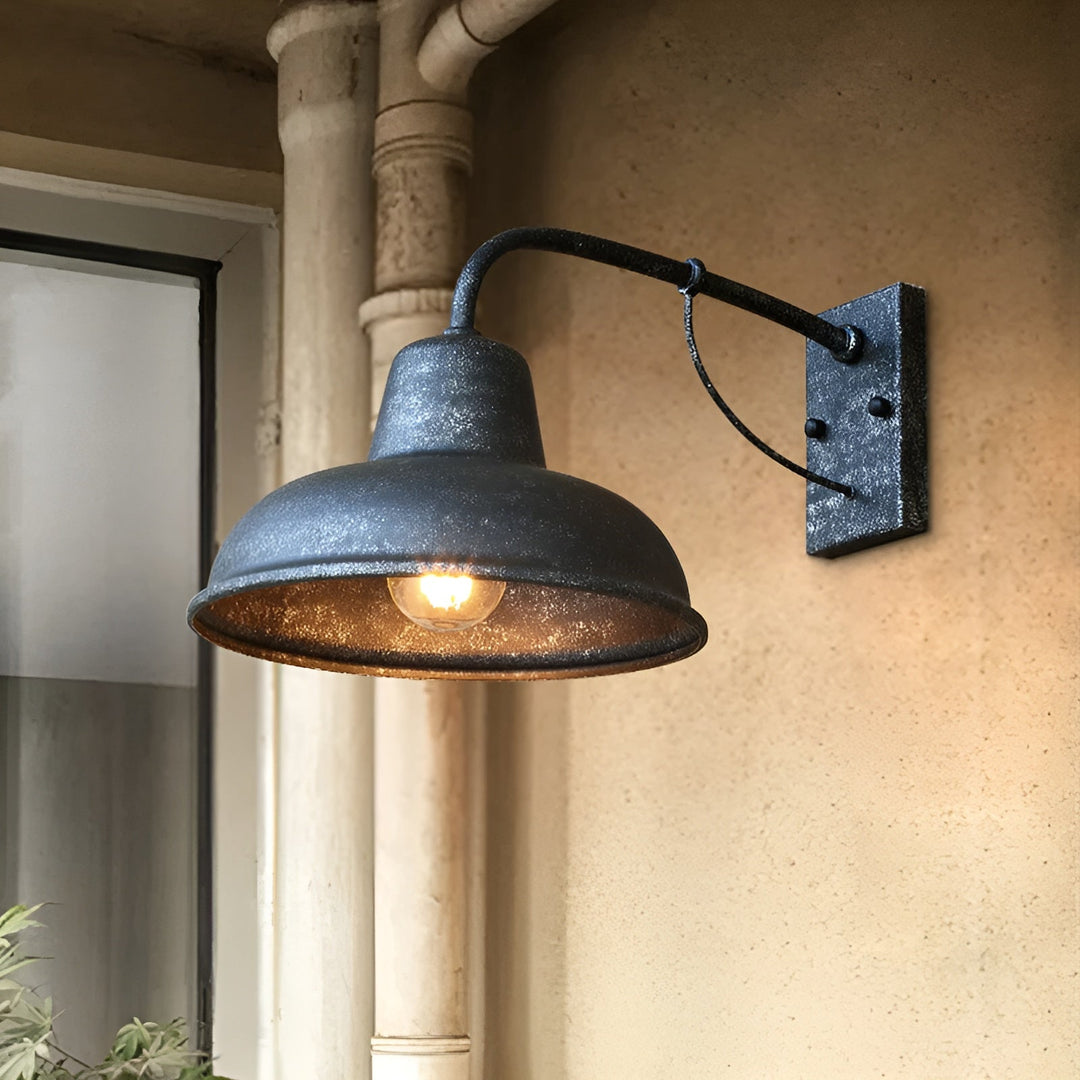 Wandlamp met verweerde donkergrijze afwerking en brandende lamp aan een zandkleurige muur naast een regenpijp en raam. 6