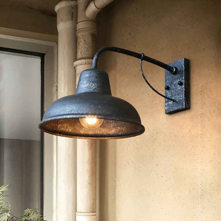 Wandlamp met verweerde donkergrijze afwerking en brandende lamp aan een zandkleurige muur naast een regenpijp en raam. 6