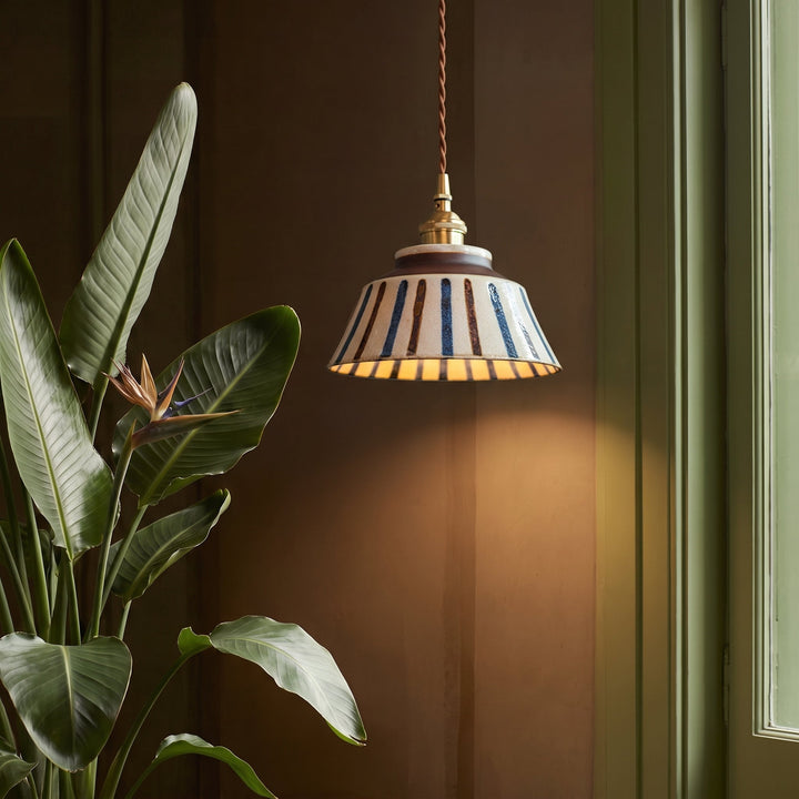 Gestreepte keramische hanglamp brandt naast een grote plant met vogelbloem, tegen een warme bruine muur en groen raamkozijn. 4